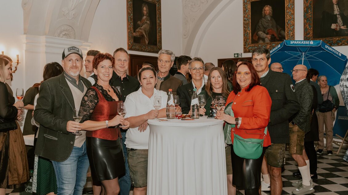 Eine Gruppe von Menschen steht um einen Stehtisch in einem festlichen Raum. Sie lächeln und halten Gläser in der Hand, umgeben von historischen Gemälden. | © cdm