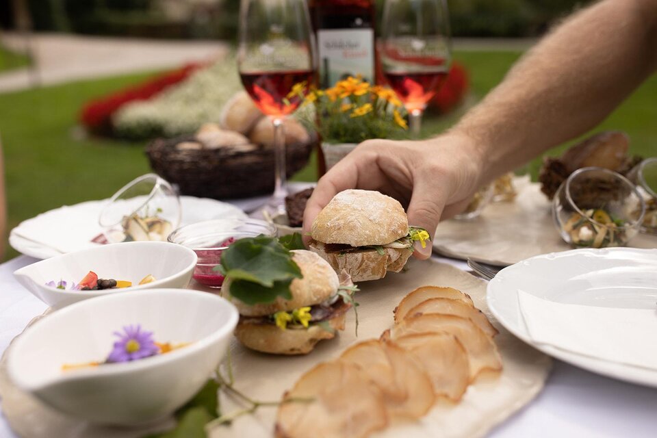 Ein rustikaler Tisch mit verschiedenen köstlichen Speisen, darunter Sandwiches mit frischen Kräutern und Aufschnitt. Im Hintergrund stehen Gläser mit Rotwein und dekorative Blumen. | © TV Südsteiermark - achromaticphotography
