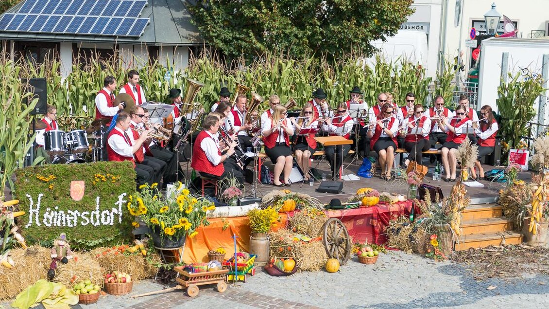 Eine Musikkapelle spielt auf einer Bühne, umgeben von herbstlichen Dekorationen. Im Hintergrund sind Sonnenblumen und Kürbisse zu sehen, die die festliche Atmosphäre unterstreichen. | © Jean Van Luelik