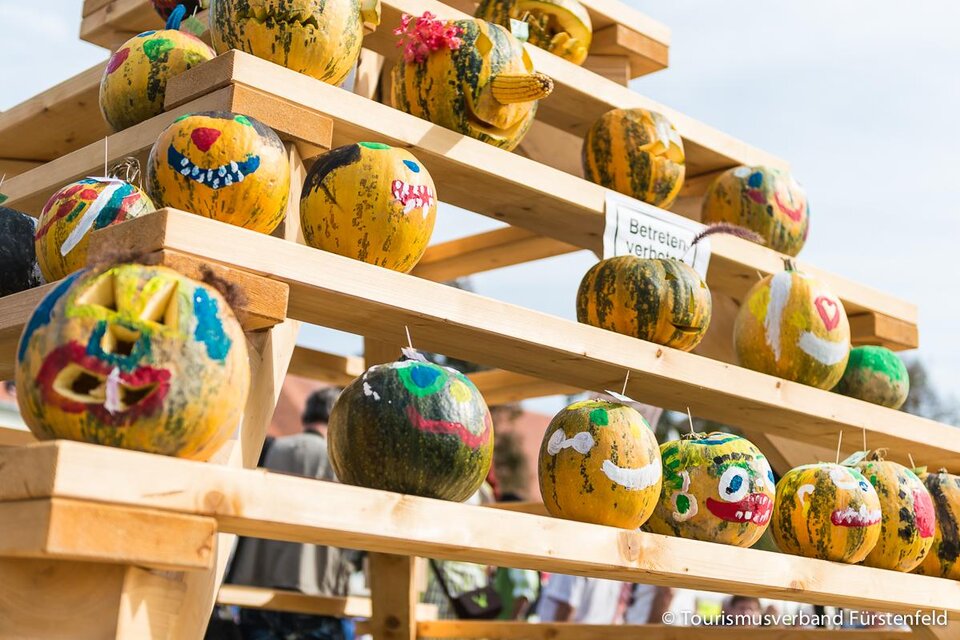 Eine Holzstruktur mit mehreren bemalten Kürbissen, die fröhliche Gesichter zeigen. Die Kürbisse sind bunt gestaltet und stehen auf verschiedenen Ebenen. | © Jean Van Luelik Photographer