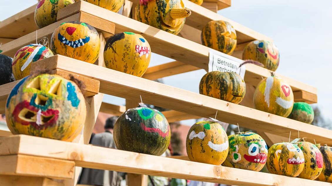Eine Holzstruktur mit mehreren bemalten Kürbissen, die fröhliche Gesichter zeigen. Die Kürbisse sind bunt gestaltet und stehen auf verschiedenen Ebenen. | © Jean Van Luelik Photographer