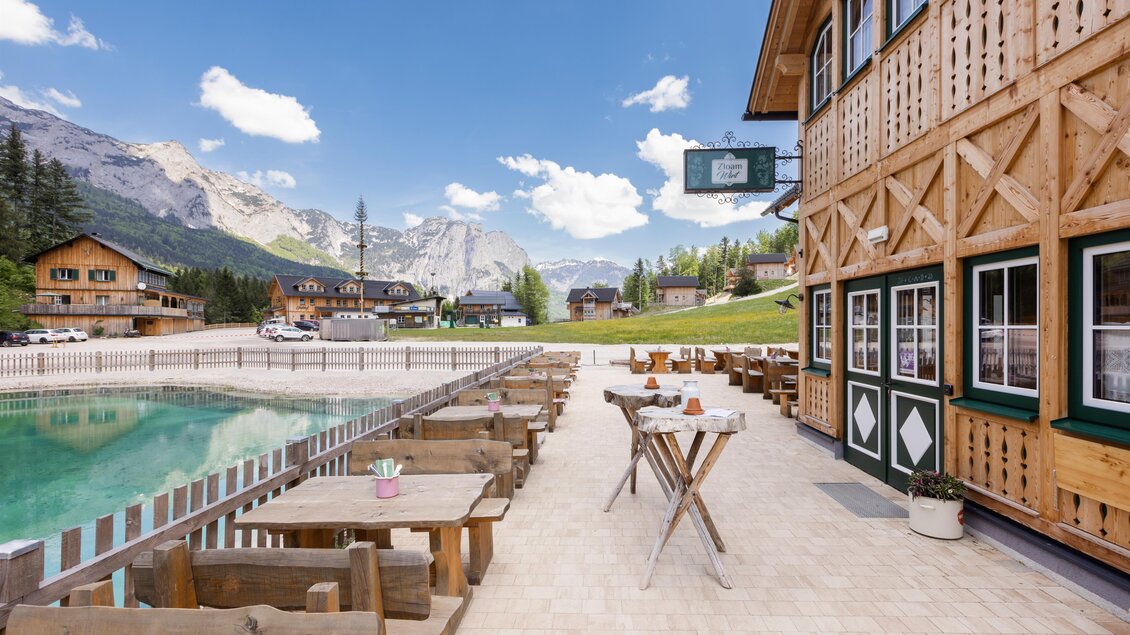 Eine alpine Landschaft mit einem klaren See und einem Holzhäusern im Hintergrund. Auf der Terrasse stehen Tische und Stühle, ideal zum Entspannen in der Natur.
