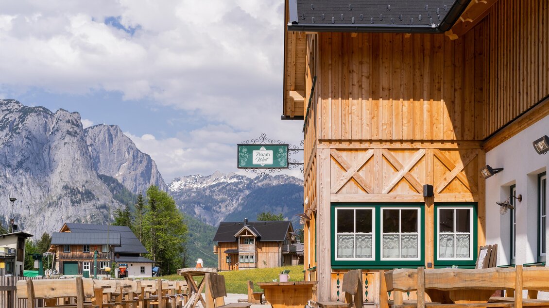 Ein rustikales Holzgebäude mit Terrasse und Blick auf die Berge. Der Himmel ist klar mit einigen Wolken. | © www.zloamwirt.at