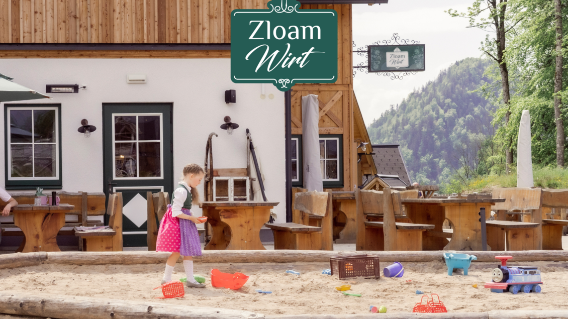 Ein kinderfreundlicher Außenbereich mit Sandkasten und Spielzeug. Im Hintergrund ein traditionelles Gasthaus umgeben von Bäumen. | © Zloam Wirt, www.zloamwirt.at