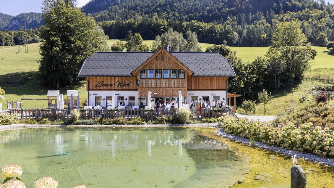 Ein charmantes Holzgebäude liegt am Ufer eines klaren Teichs, umgeben von grünen Wiesen und sanften Hügeln. Im Hintergrund erheben sich majestätische Berge unter einem blauen Himmel. | © www.zloamwirt.at