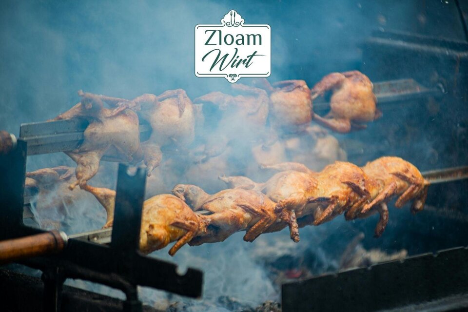 Gegrillte Hähnchen, die über offenem Feuer auf Spießen zubereitet werden. Rauch zieht durch die Szene und bringt eine einladende Atmosphäre. | © www.zloamwirt.at