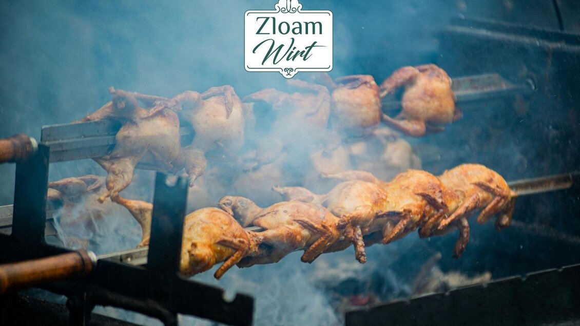 Gegrillte Hähnchen, die über offenem Feuer auf Spießen zubereitet werden. Rauch zieht durch die Szene und bringt eine einladende Atmosphäre. | © www.zloamwirt.at