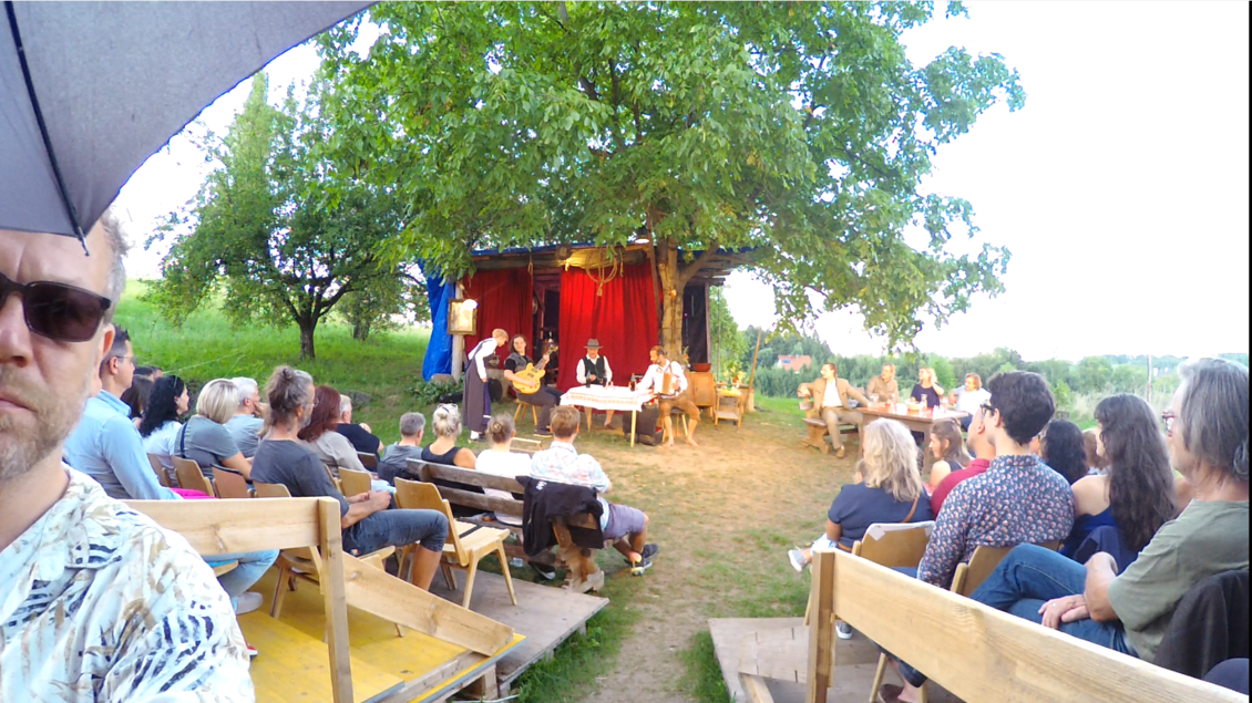Eine Freiluftveranstaltung mit einem Theaterstück unter einem Baum. Das Publikum sitzt auf Bänken und schaut den Darstellern auf der Bühne zu. | © Jimi Lend