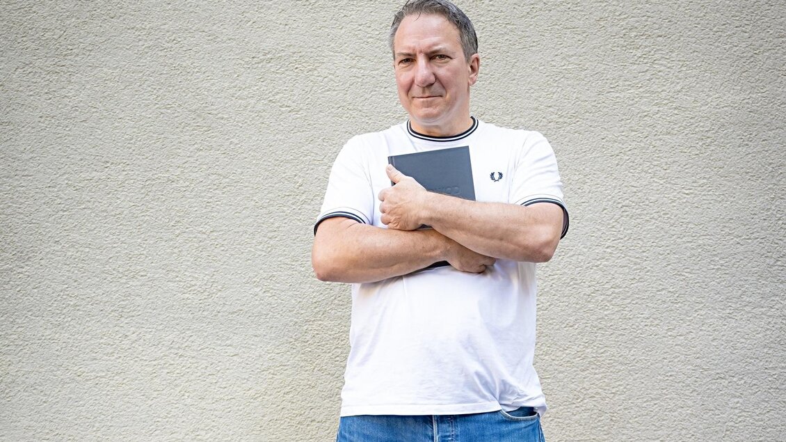 Robert Palfrader steht mit seinem Buch am Gehsteig an einer Hauswand  | © wirlphoto