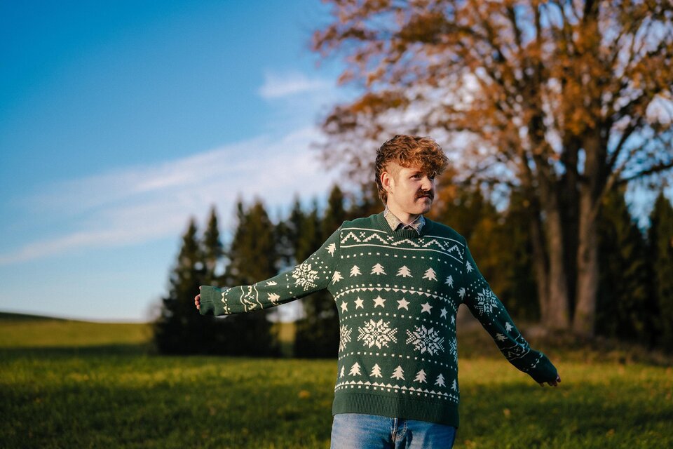 Ein Mann in einem grünen Weihnachtspullover steht in einem Feld. Im Hintergrund sind Bäume und ein blauer Himmel zu sehen. | ©  Anna Sophie Koelbl