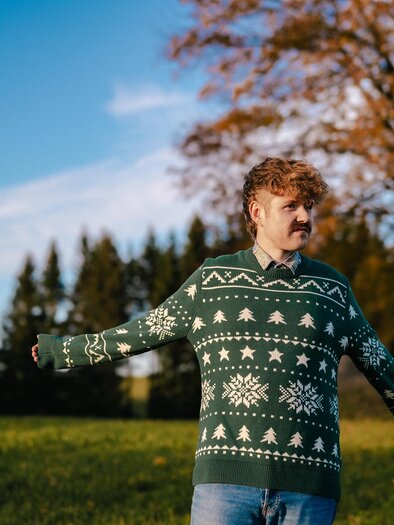 A man in a green Christmas sweater stands in a field. In the background, there are trees and a blue sky. | ©  Anna Sophie Koelbl
