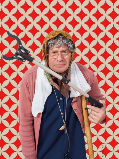 An older woman with glasses stands in front of a colorful, patterned background. She is holding a cane and is wearing a headpiece. | ©  Christopher Marvic
