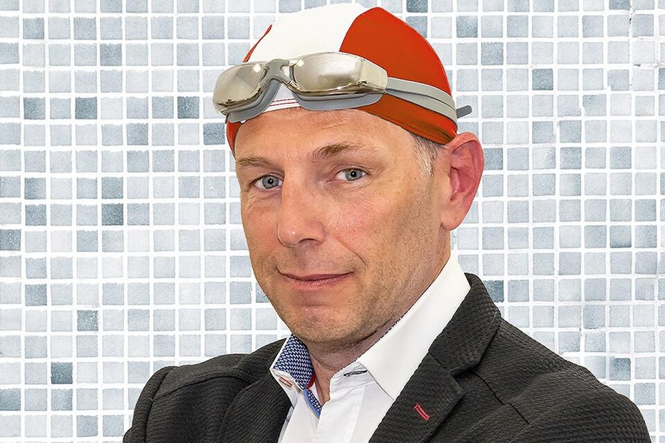 Ein Mann in einem Schwimmkostüm mit einer roten Badekappe und Schwimmbrille. Er steht vor einer Mosaikwand und sieht selbstbewusst aus. | ©  Rudi Ferder