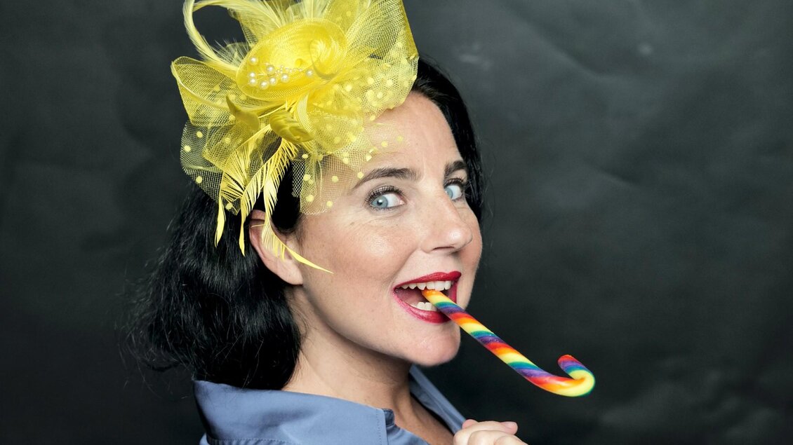Kabarettistin Nadja Maleh mit auffälligem gelbem Fascinator und buntem Lutscher vor dunklem Hintergrund. | © Sabine Stieger