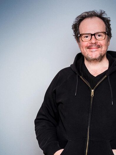A smiling man with glasses is wearing a black hoodie. The background is in soft shades of gray. | © Thomas Mraz