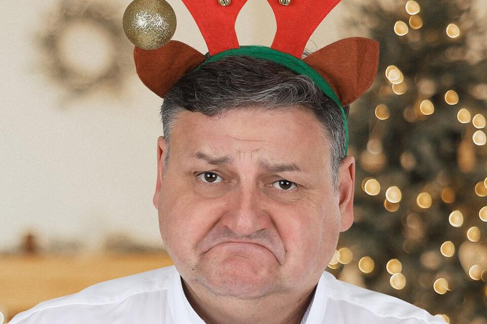 Ein Mann mit einem traurigen Gesicht trägt ein Rentiergeweih. Im Hintergrund sind Weihnachtslichter und dekorierte Bäume zu sehen. | © Gerhard Sengstschmid