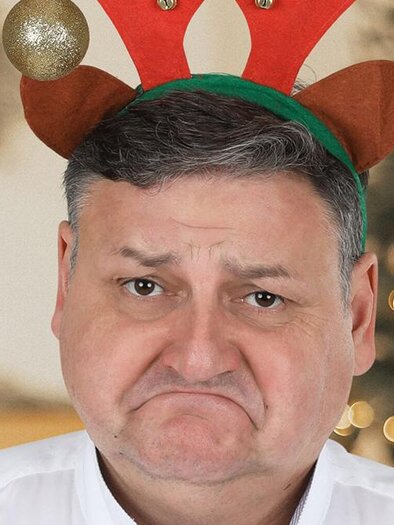 Ein Mann mit einem traurigen Gesicht trägt ein Rentiergeweih. Im Hintergrund sind Weihnachtslichter und dekorierte Bäume zu sehen. | © Gerhard Sengstschmid