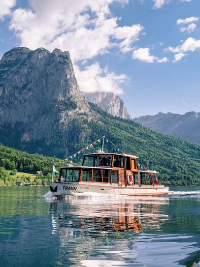 A boat glides quietly over a sparkling lake, surrounded by majestic mountains. The sky is clear with some white clouds. | © Schifffahrt Grundlsee Toplitzsee