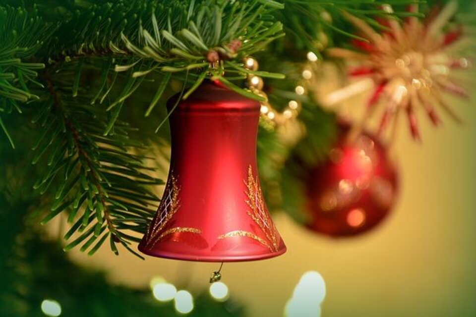 Eine rote Weihnachtsglocke hängt an einem Tannenzweig. Im Hintergrund sind weitere festliche Dekorationen und Lichter sichtbar. | © Pixabay