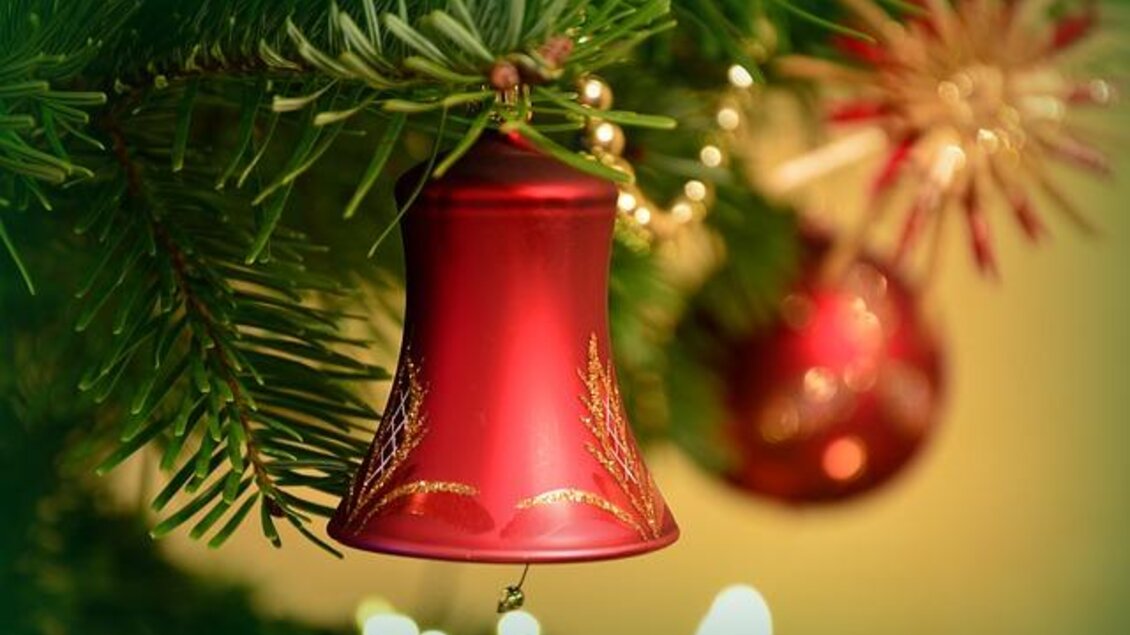 Eine rote Weihnachtsglocke hängt an einem Tannenzweig. Im Hintergrund sind weitere festliche Dekorationen und Lichter sichtbar. | © Pixabay