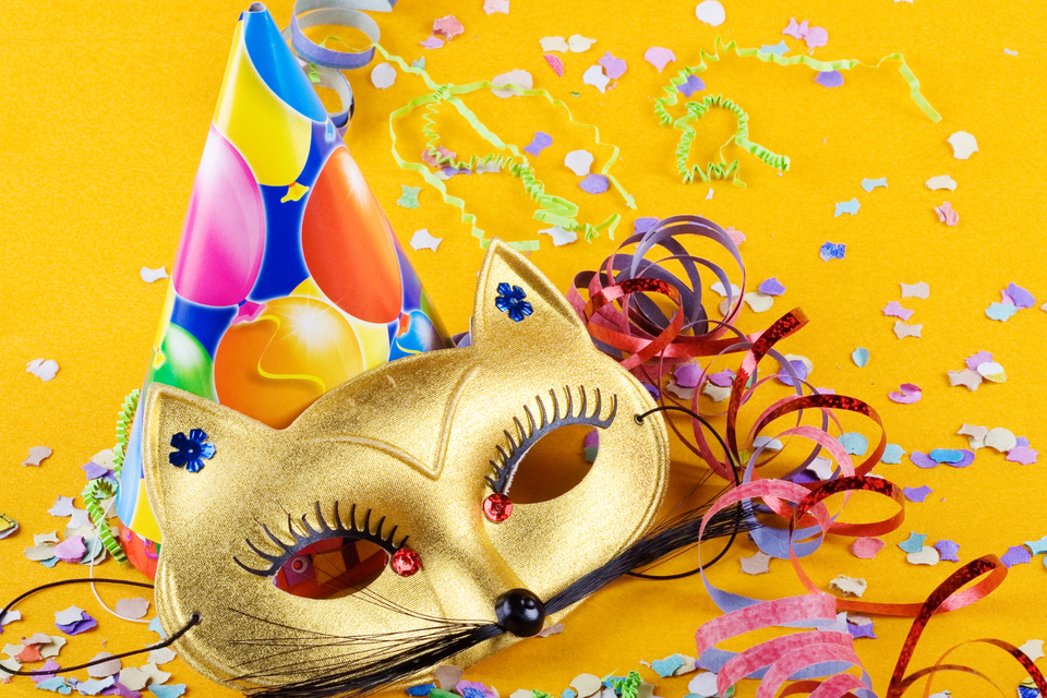Eine goldene Faschingsmaske mit Schnurrhaaren und Katzenohren, umgeben von buntem Konfetti und verschieden gemusterten, bunten Luftschlangen und einem Partyhut mit bunten Luftballons liegen auf orangefarbenem Hintergrund. | © Canva