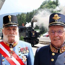 Two men in historical uniforms stand in front of a steam-powered locomotive. In the background, green hills and a blue sky are visible.