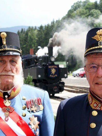 Two men in historical uniforms stand in front of a steam-powered locomotive. In the background, green hills and a blue sky are visible.