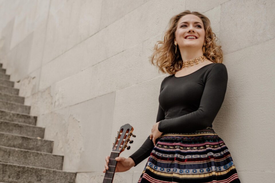 Junge Frau mit Gitarre in der Hand, die an einer Wand lehnt und lächelt. Sie trägt ein schwarzes Oberteil und einen bunten Rock, während ihre Haare leicht im Wind wehen. Im Hintergrund sind Treppenstufen zu sehen. | © Horacio Reyes Paez