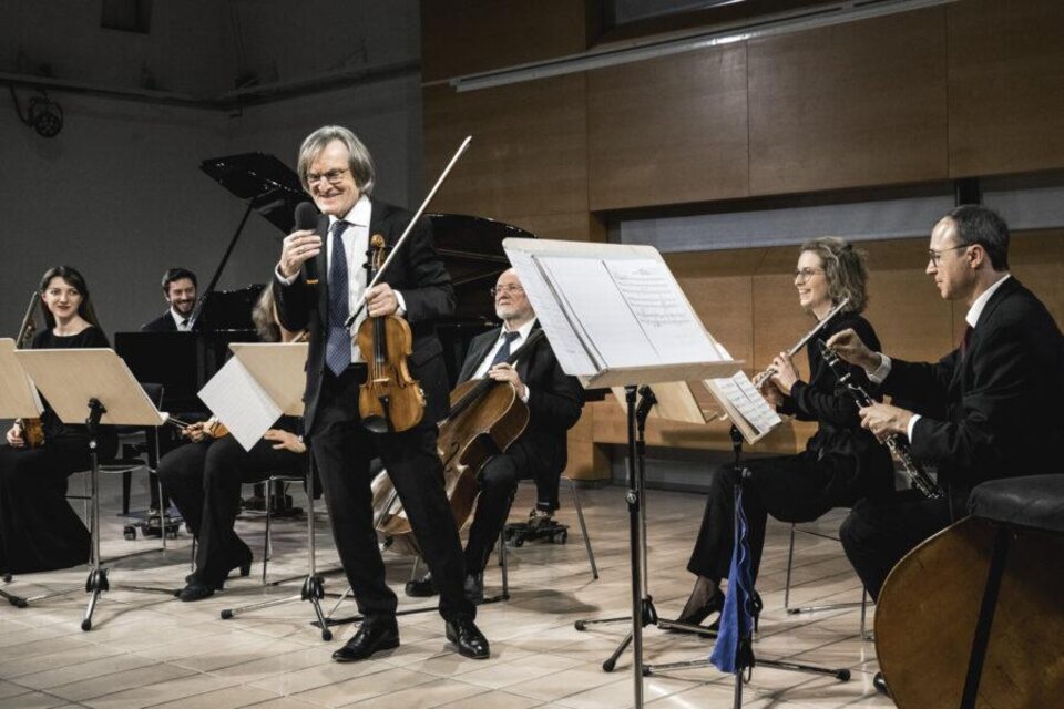 Ein Musiker mit einer Violine steht im Vordergrund und spricht lächelnd zu seinem Publikum. Im Hintergrund sind weitere Musiker mit verschiedenen Instrumenten zu sehen, die in einem modernen Raum auftreten. | © Ulrike Rauch