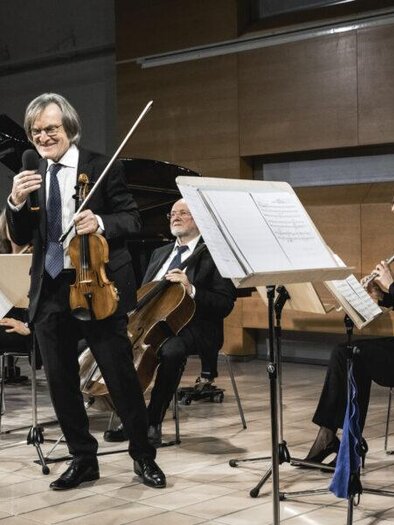 Ein Musiker mit einer Violine steht im Vordergrund und spricht lächelnd zu seinem Publikum. Im Hintergrund sind weitere Musiker mit verschiedenen Instrumenten zu sehen, die in einem modernen Raum auftreten. | © Ulrike Rauch