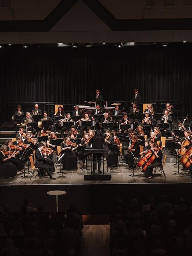 One sees the impressive size of the Gleisdorf Chamber Orchestra under the direction of Ms. Yu-Hao Lorenz, who stands in the middle as the conductor, in front of more than fifty musicians elegantly dressed in black, with various instruments - from violins and cellos to flutes, trumpets, and other wind instruments as well as percussion instruments in the background. | © Oststeiermark Tourismus