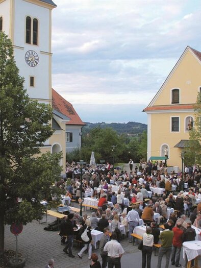 Ein lebendiger Platz mit vielen Menschen, die an Tischen sitzen. Im Hintergrund sind eine Kirche und historische Gebäude zu sehen. | © TV Südsteiermark