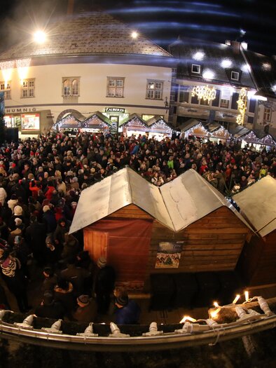 A bustling Christmas market at night with many people and festive lighting. Stalls with traditional wooden huts can be seen in the foreground.