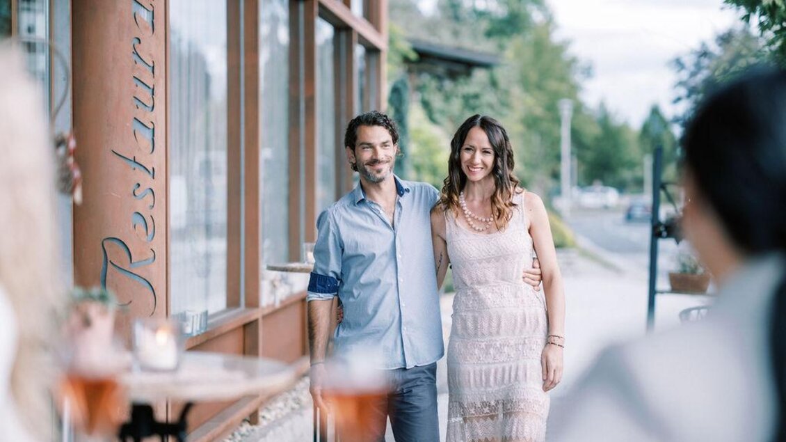 Ein Paar spaziert lächelnd vor einem Restaurant. Die Frau trägt ein elegantes Kleid, während der Mann in lässiger Kleidung erscheint. | © Garten Hotel Ochensberger
