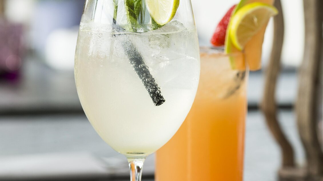 Zwei farbenfrohe Cocktails stehen auf einem Tisch. Der erste ist ein heller Cocktail mit Limetten und Minze, der zweite ist orangefarben mit einer Limettenscheibe. | © Garten Hotel Ochensberger