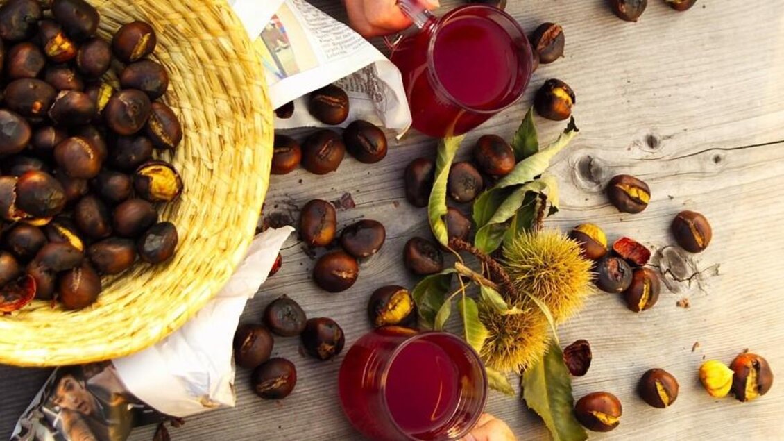 Geröstete Kastanien, Trauben und ein Glas roter Saft auf einem Holzbrett. Die Farben sind warm und einladend. | © © Werner Krug