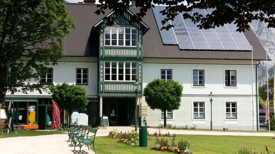 Ein historisches Gebäude in Altaussee mit grünen Akzenten und Solarpanelen auf dem Dach. Vor dem Haus befindet sich ein gepflegter Garten mit bunten Blumen und Bänken. | © Petra Kirchschlager