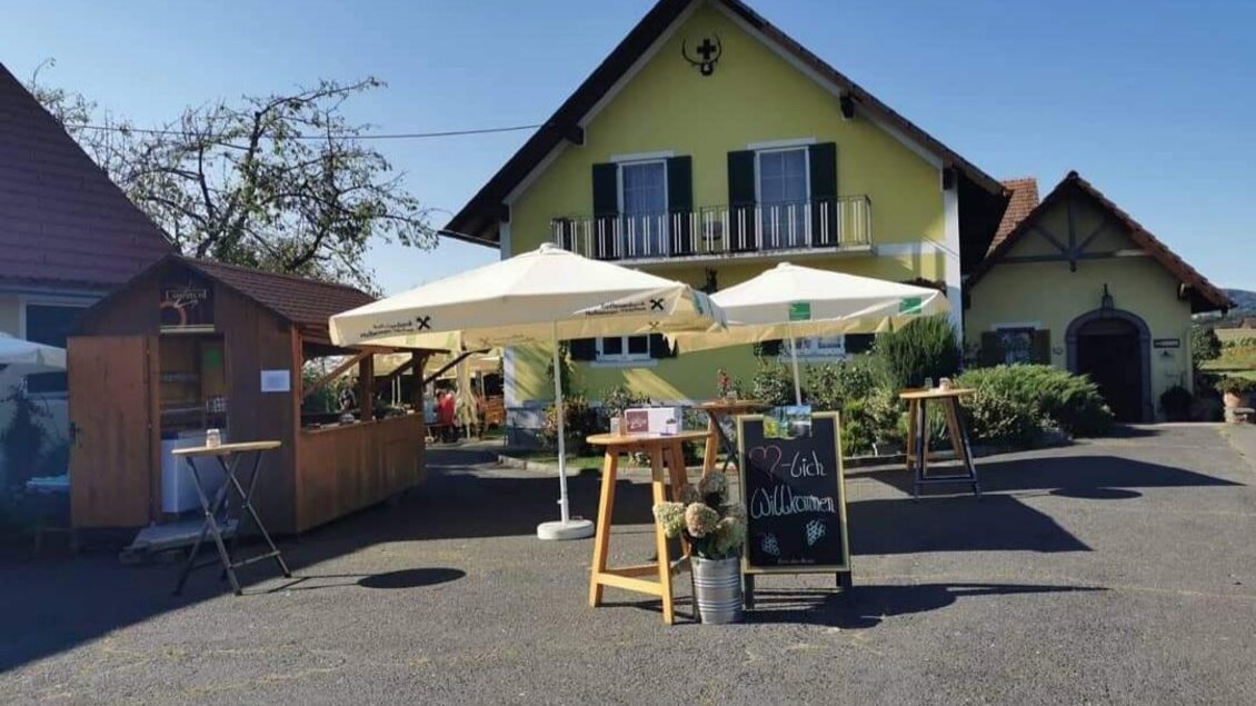Ein gelbes Gebäude mit einem begrünten Außenbereich. Pavillons und Tische sind auf einem gepflasterten Platz aufgestellt. | © Weinbau Lamprecht