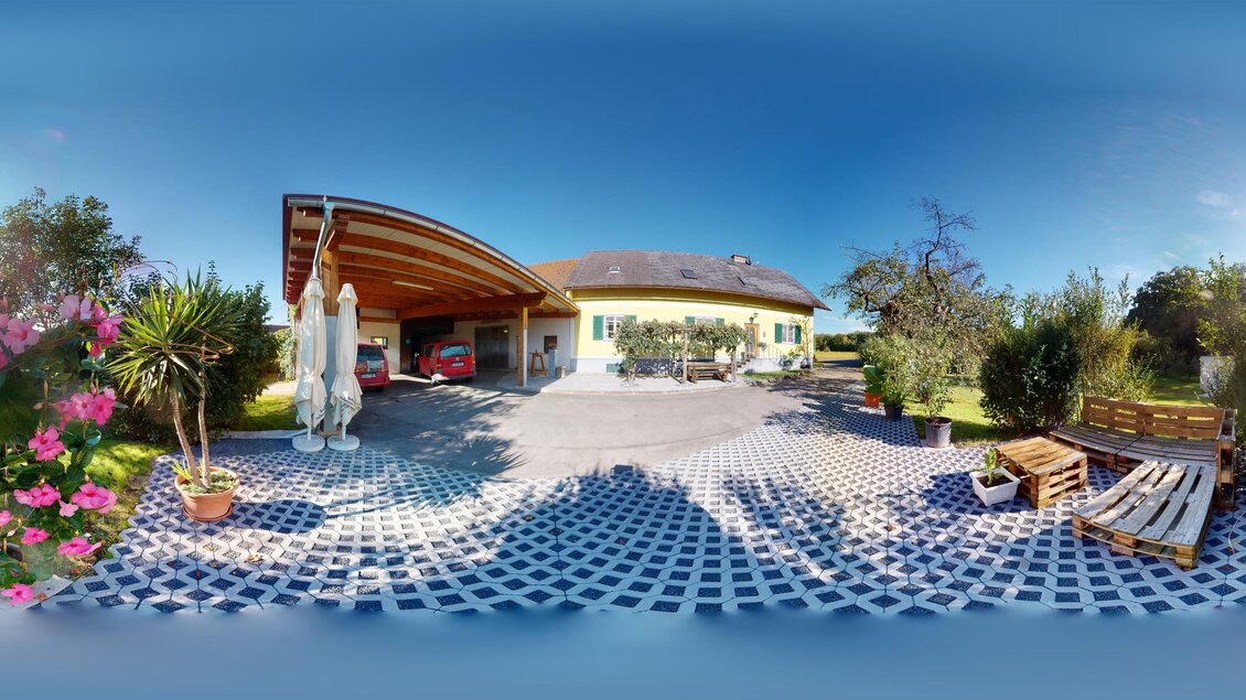 Ein schönes Grundstück mit einem modernen Haus und einer gepflasterten Einfahrt. Blumen und Palmen schmücken den Außenbereich unter einem klaren blauen Himmel. | © Weinbau Lamprecht