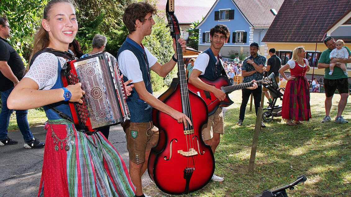 Eine Gruppe junger Musiker spielt traditionelle Instrumente im Freien. Im Hintergrund sind Menschen bei einer Veranstaltung zu sehen. | © Helmut Adelwöhrer