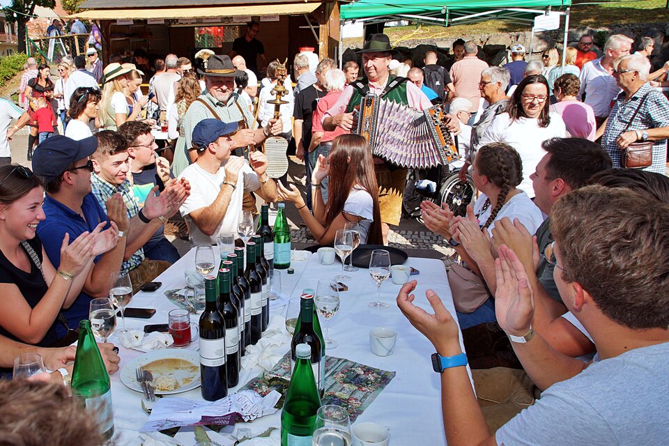 Eine lebhafte Feier mit vielen Menschen, die an einem Tisch sitzen und applaudieren. Ein Musiker spielt Akkordeon und sorgt für gute Stimmung. | © Helmut Adelwöhrer