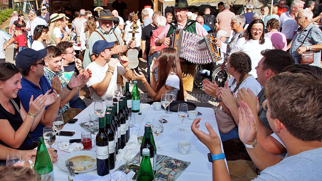 Eine lebhafte Feier mit vielen Menschen, die an einem Tisch sitzen und applaudieren. Ein Musiker spielt Akkordeon und sorgt für gute Stimmung. | © Helmut Adelwöhrer