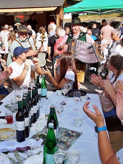 Eine lebhafte Feier mit vielen Menschen, die an einem Tisch sitzen und applaudieren. Ein Musiker spielt Akkordeon und sorgt für gute Stimmung. | © Helmut Adelwöhrer