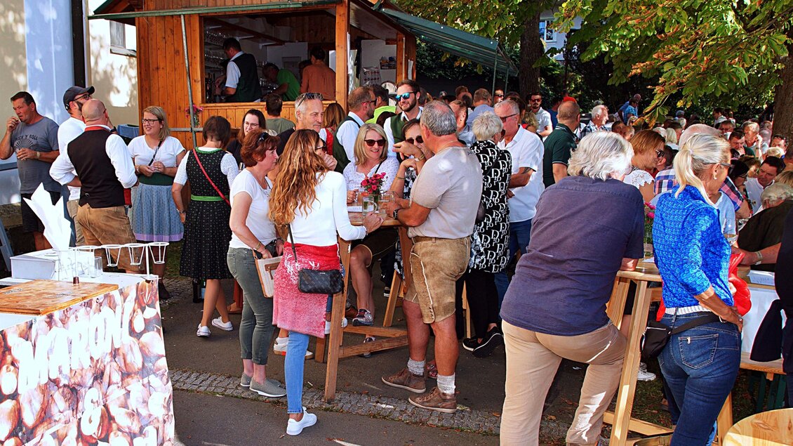Eine belebte Veranstaltung im Freien mit vielen Menschen, die an einem Stand stehen und sich unterhalten. Im Hintergrund ist ein Holzstand und Bäume zu sehen. | © Helmut Adelwöhrer