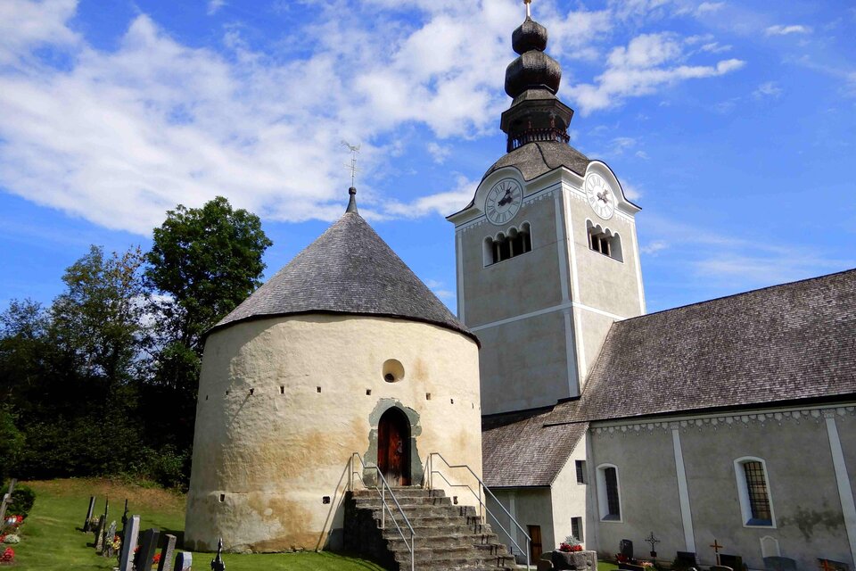 Eine historische Kirche mit einer runden Kapelle und einem hohen Turm. Umgeben von grünem Rasen und Grabsteinen im Vordergrund.