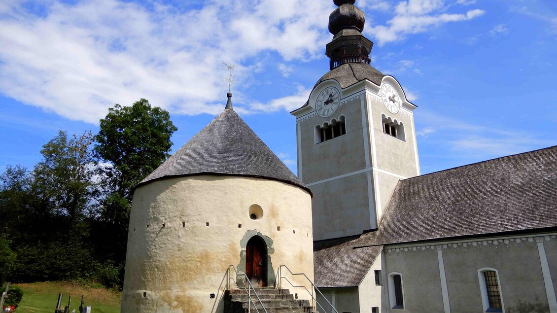 Eine historische Kirche mit einer runden Kapelle und einem hohen Turm. Umgeben von grünem Rasen und Grabsteinen im Vordergrund.
