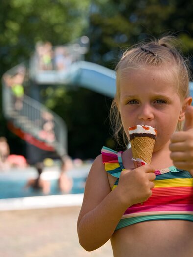 Ein kleines Mädchen mit einem Eis in der Hand zeigt den Daumen nach oben. Im Hintergrund sind ein Schwimmbecken und eine Rutsche zu sehen. | © Motion Manager