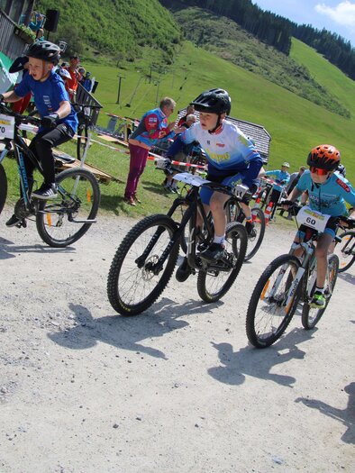 Eine Gruppe von Kindern fährt begeistert mit dem Mountainbike auf einem Schotterweg. Im Hintergrund sind grüne Hügel und Zuschauer zu sehen. | © Johann Schröcker