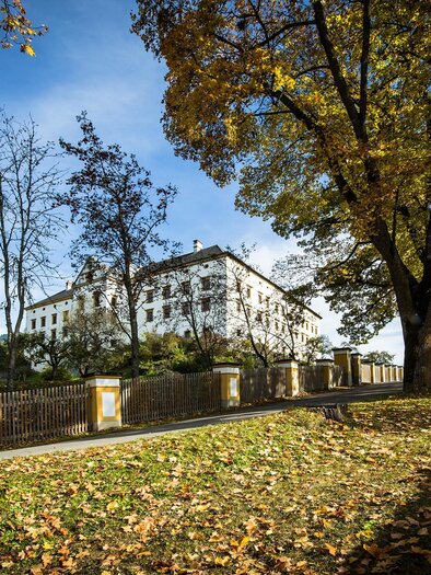 Ein historisches Gebäude umgeben von herbstlichen Bäumen. Der Weg ist mit buntem Laub bedeckt.