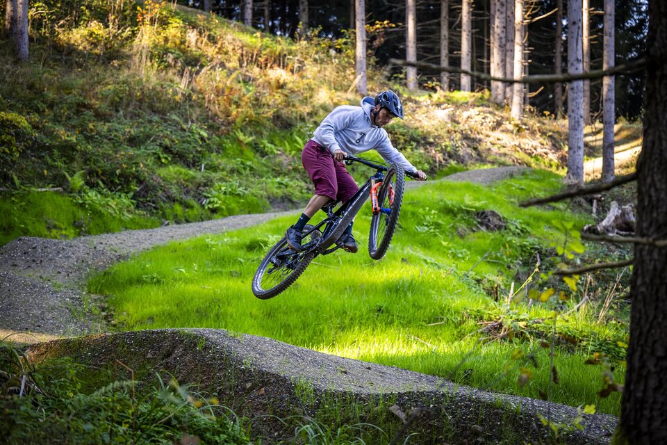 Ein Radfahrer springt über eine Erhebung in einem Waldgebiet. Die Umgebung ist grün und von Bäumen umgeben. | © Lucas Pripfl